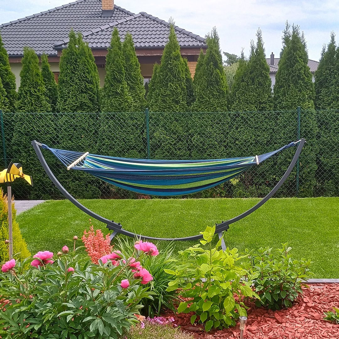 Hängematte mit Metallgestell auf grünem Rasen vor dicht bepflanzter Hecke und Blumenbeet