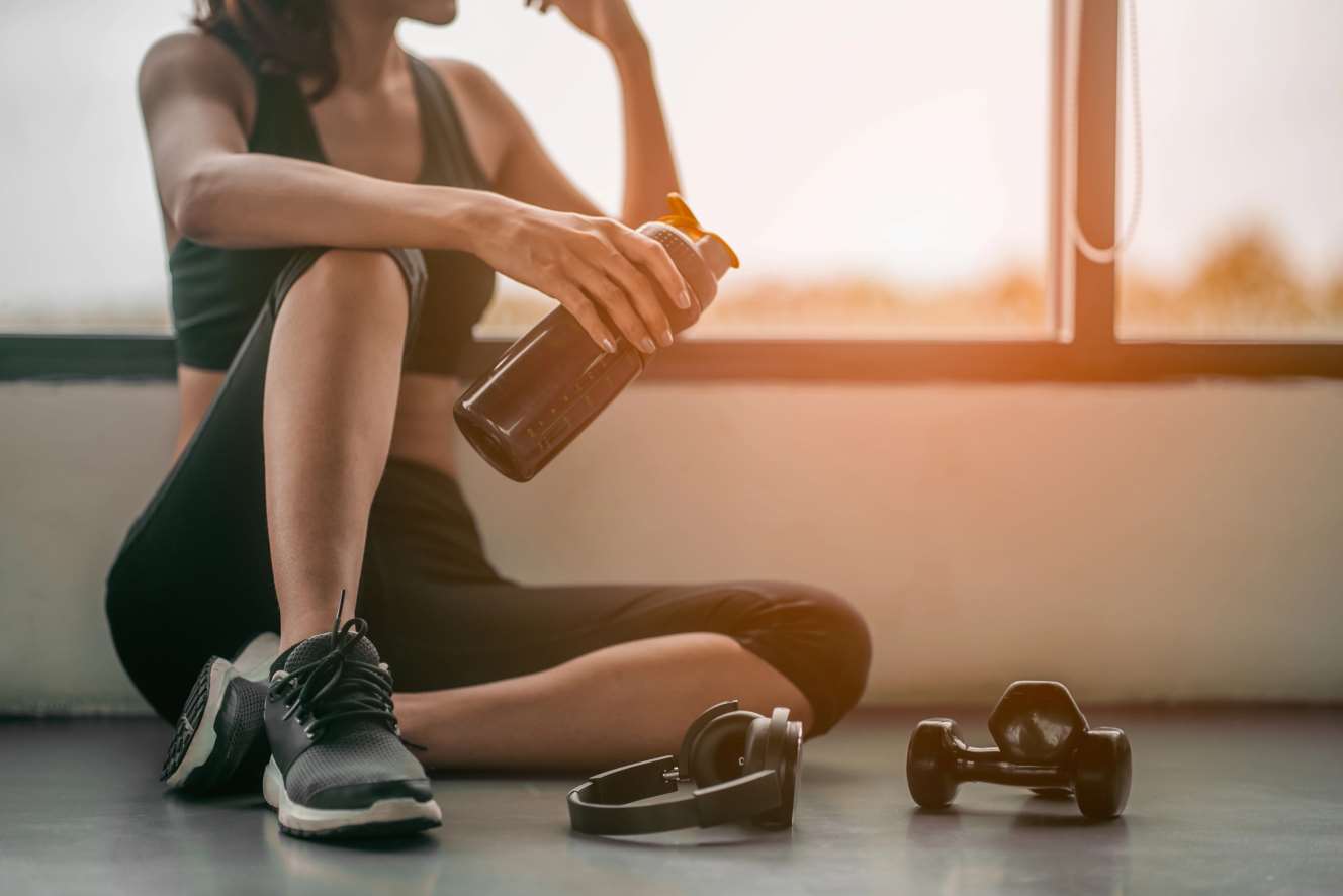 Eine Frau im Fitnessstudio entspannt sich nach dem Training, im Vordergrund sind eine Wasserflasche, Hanteln und Kopfhörer zu sehen.