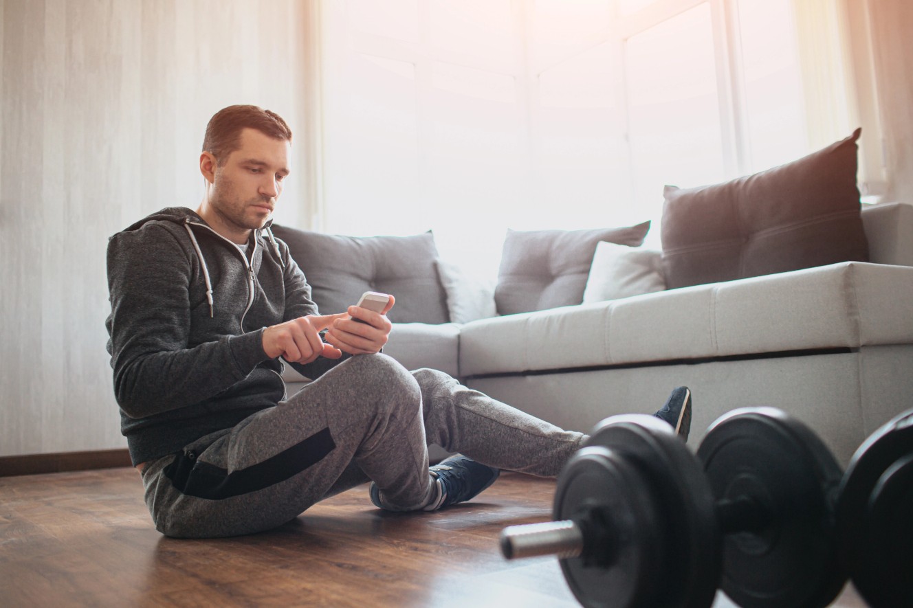 Mann sitzt vor Hanteln und schaut auf sein Smartphone – typische Ablenkung beim Training zu Hause